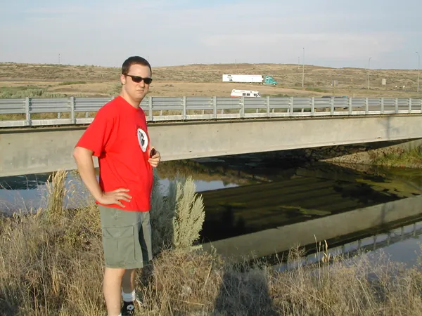 A traveler poses at a roadside overlook above a bridge.